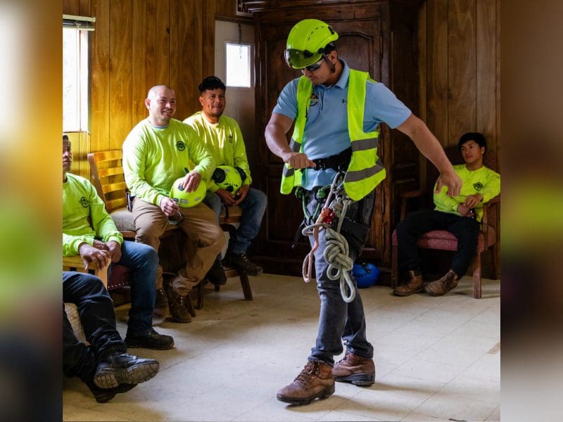 A person demonstrates climbing gear to attentive onlookers in a rustic room.